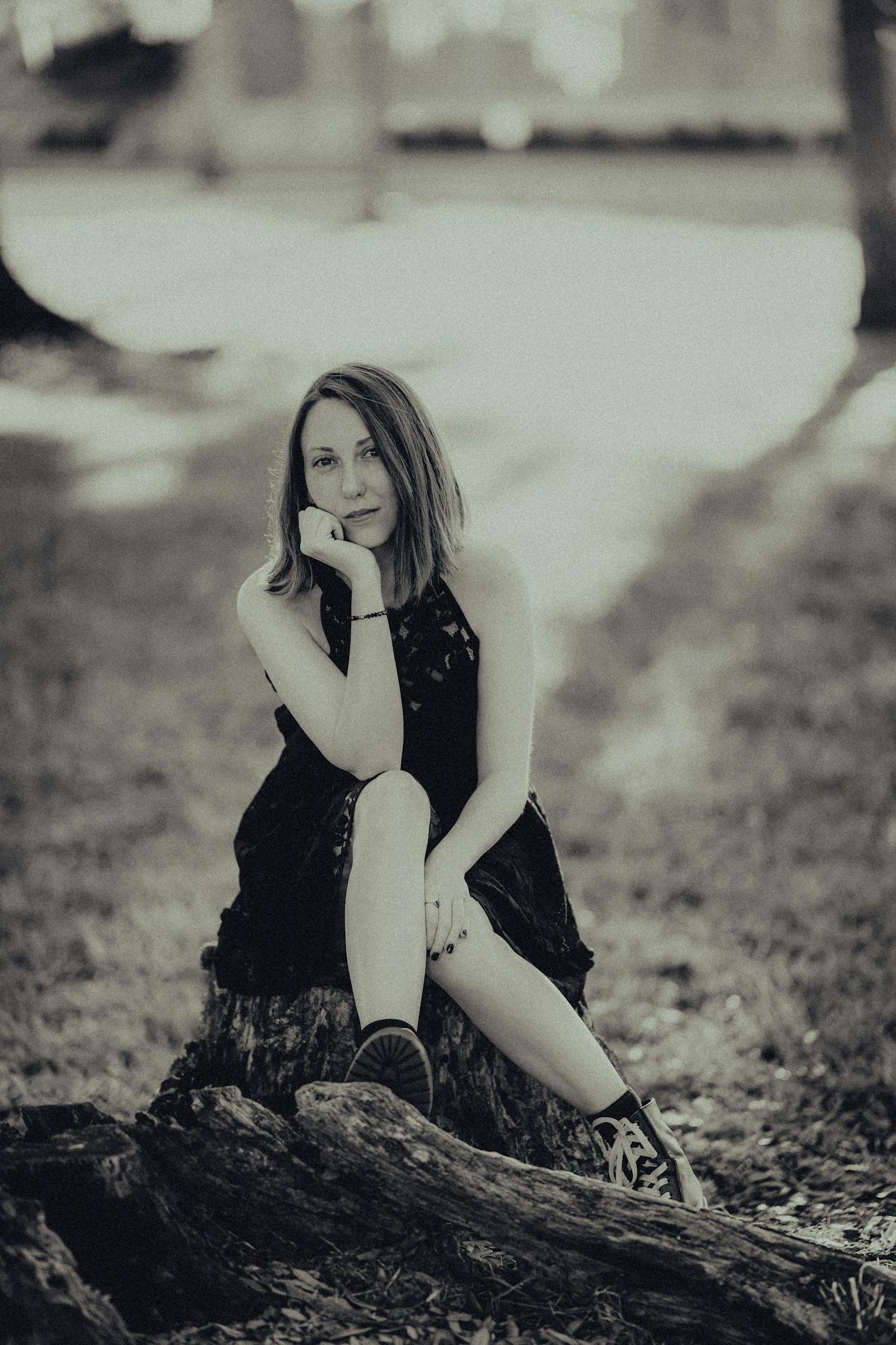 A woman in a black dress sits contemplatively on a tree stump in a tranquil park.